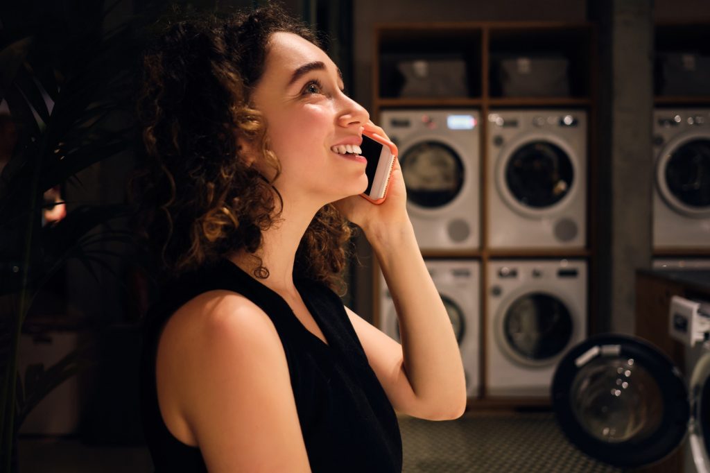 Pretty girl happily talking on smartphone waiting washing in modern self-service laundry at night